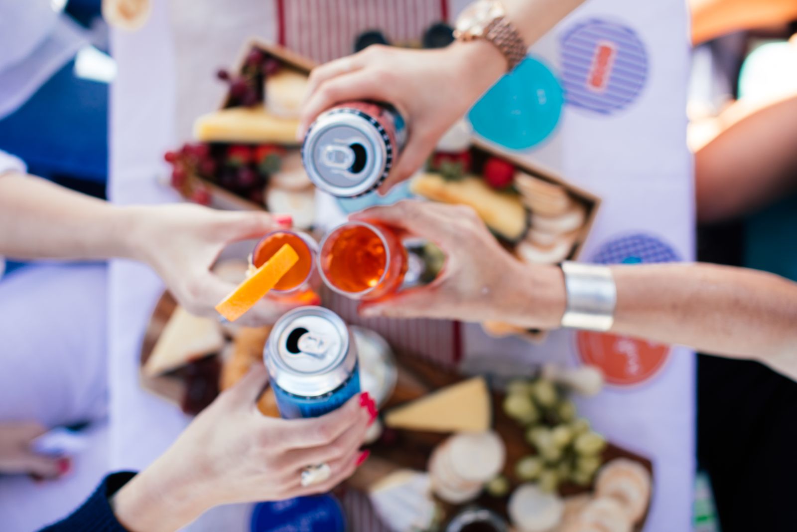 Crop anonymous people clinking glasses and cans over table while having party in sunny day
